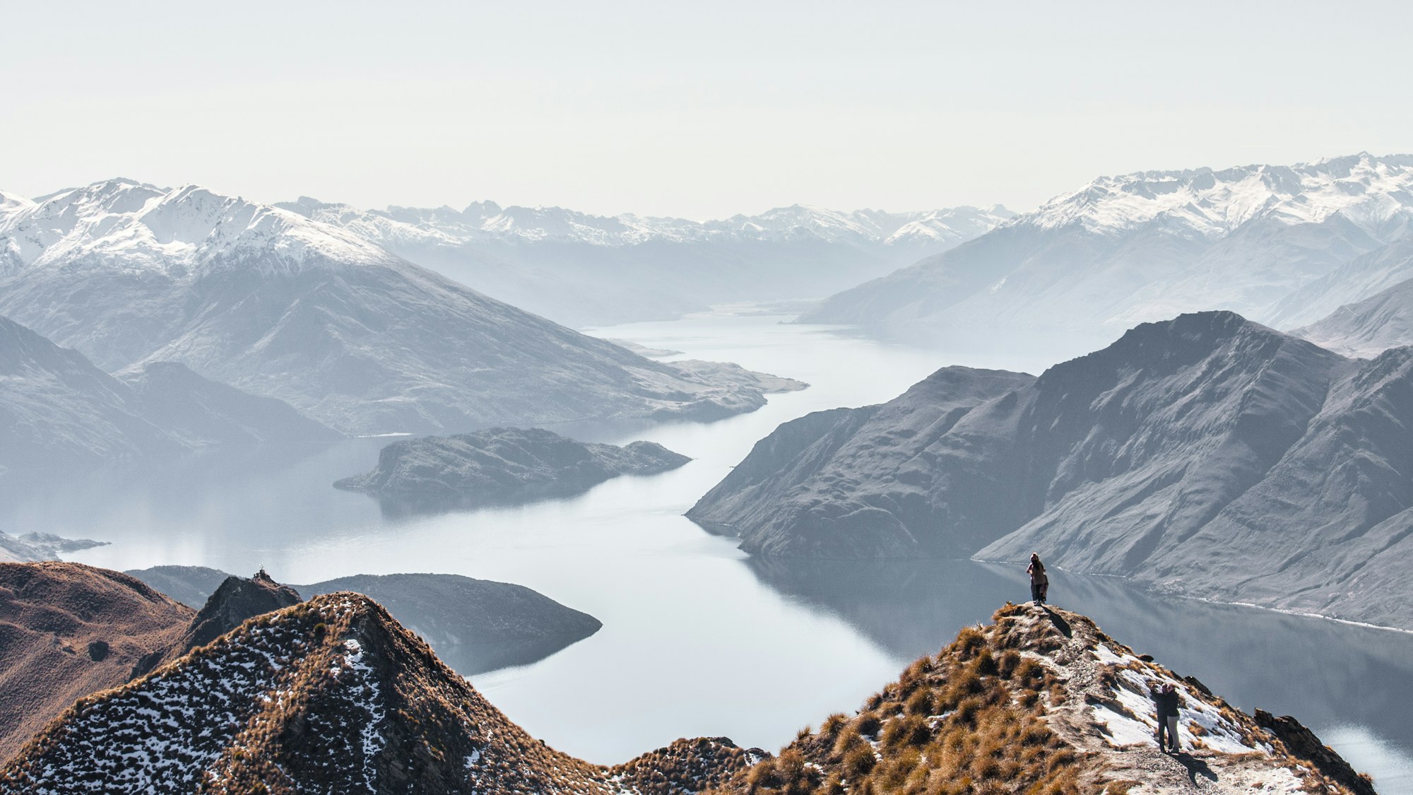 A hiker on a summit above a misty New Zealand lake and mountains
